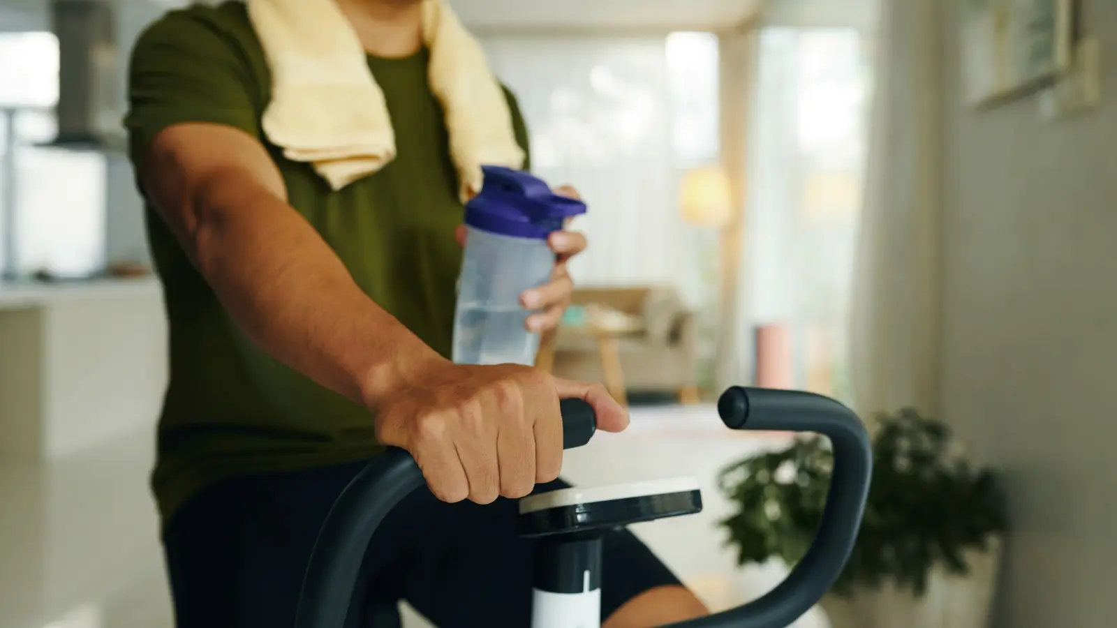 Persona che si idrata con una bottiglia d'acqua mentre fa esercizio su una cyclette a casa. Allenamento cardio domestico e recupero.
